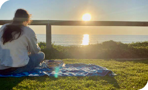 Young lady sits on a picnic blanket on the grass by the beach looking at the sunset